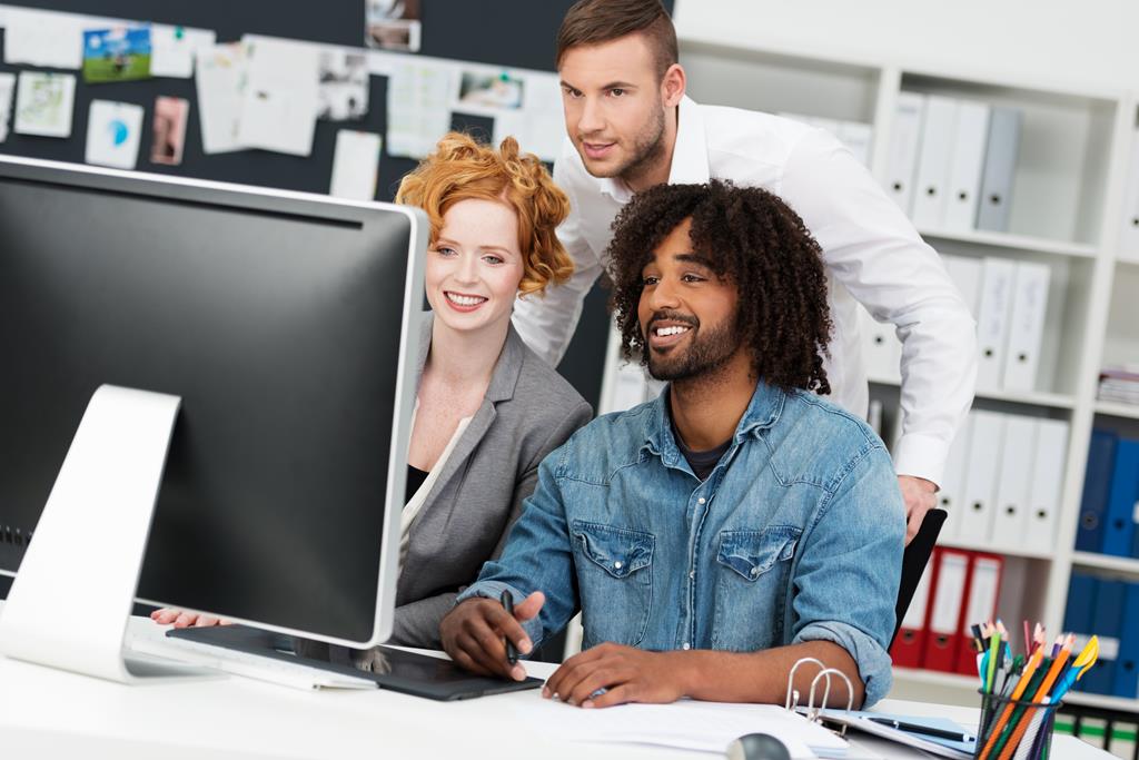 Group of diverse multiethnic business colleagues in the office smiling as they stand looking at a large desktop monitor